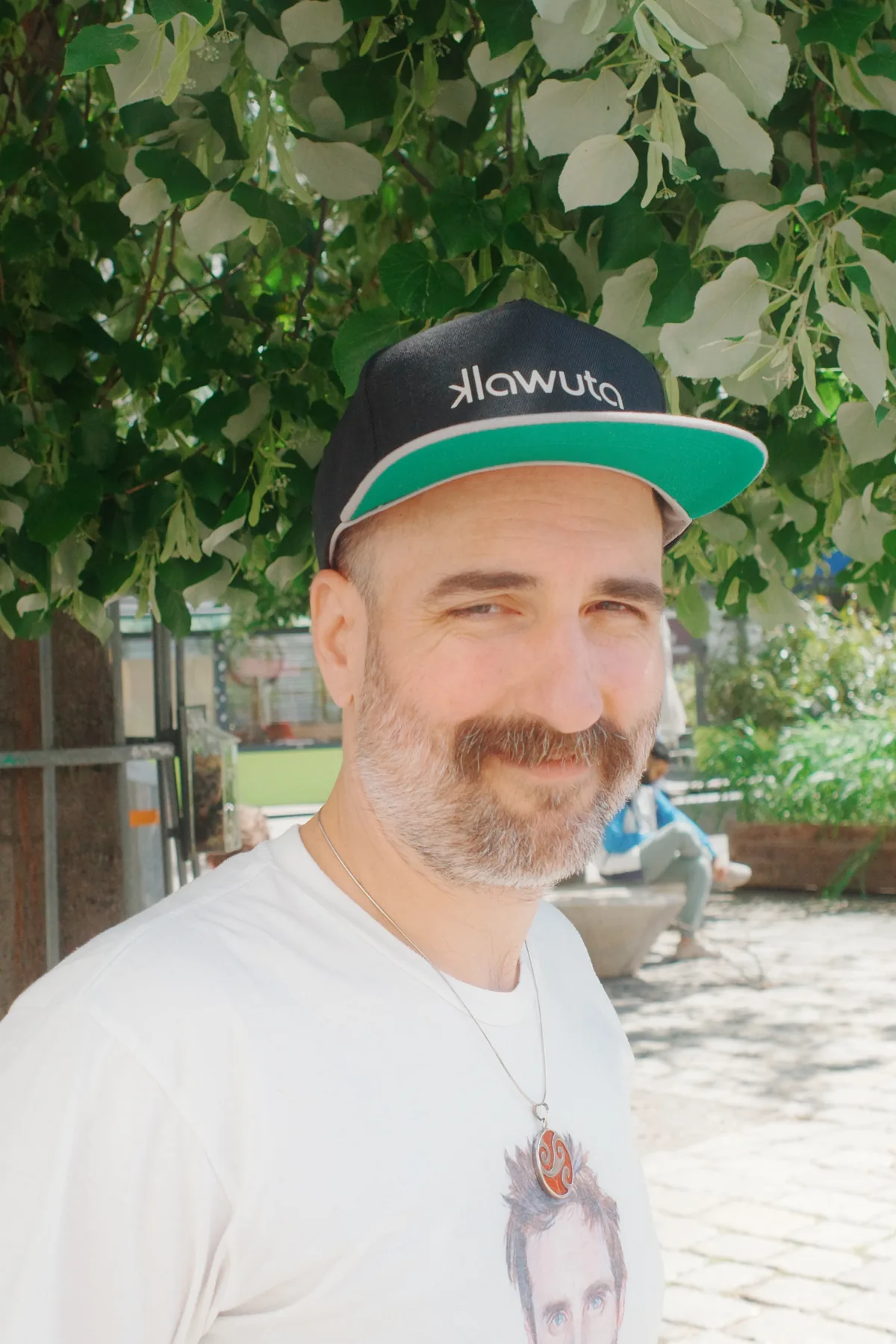Alexander Lausch im Freien unter einem Baum, mit Kappe und hellem T-Shirt, halb zur Kamera gedreht und lächelnd.